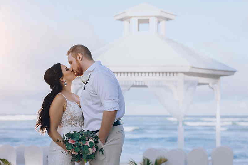 Pareja de novios besándose frente a un altar en la playa en Majestic Colonial Punta Cana
